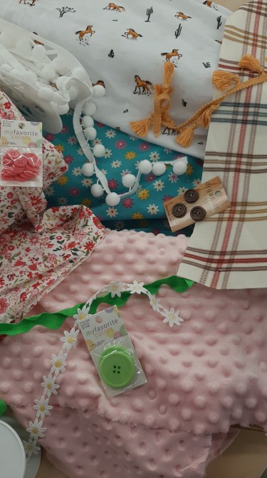 Assorted retro fabrics, like a horse print, beige/rust plaid, blue with daisies, ditsy flowers in pink, and a pink raised-dot chenille. Notions like white pom-poms, green ric rac, white daisies, gold tassels, and old buttons.